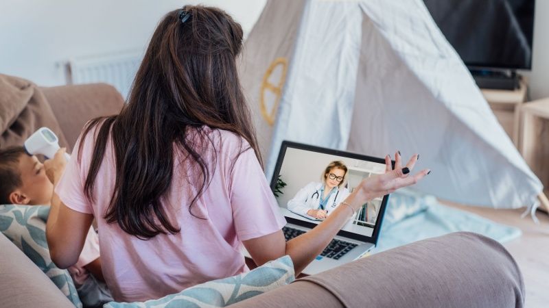 Young mother measures temperature of the child with non-contact digital infrared thermometer during online video chat with pediatrician doctor on pc.